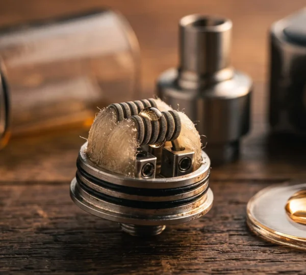A close-up of a disassembled vape atomizer showing the heating coil, cotton wick and a drop of e-liquid on a workbench.